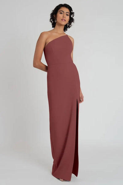 A woman in an elegant knit crepe Aubrey bridesmaid dress by Jenny Yoo with a one-shoulder neckline, posing against a neutral background.
