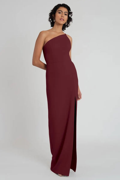 A woman in a Bergamot Bridal Jenny Yoo maroon one-shoulder evening gown stands against a neutral background.