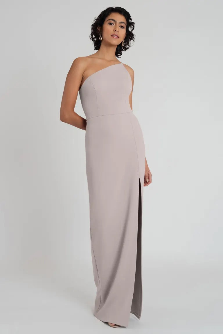 Woman posing in an elegant, Aubrey bridesmaid dress by Jenny Yoo featuring a one-shoulder neckline and made from knit crepe with a side slit from Bergamot Bridal.