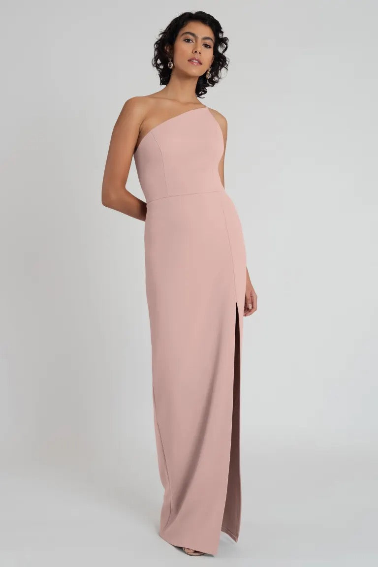 A woman poses in a pink knit crepe Aubrey bridesmaid dress by Jenny Yoo with a one-shoulder neckline and a side slit from Bergamot Bridal.