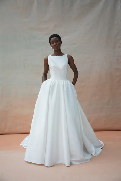 A woman stands in front of a neutral background wearing the Audrey Wedding Dress by Jenny Yoo, offered by Bergamot Bridal, which features an elegant boat neckline and a dramatic ball skirt with pockets.