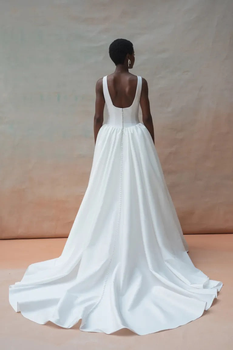 A woman in a white Audrey Wedding Dress by Jenny Yoo from Bergamot Bridal, featuring an elegant boat neckline and a dramatic ball skirt, stands facing away against a neutral backdrop.