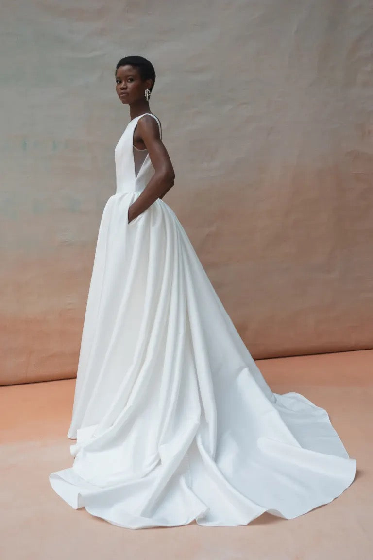 A person stands in profile wearing the Audrey Wedding Dress by Jenny Yoo from Bergamot Bridal, featuring a white, sleeveless gown with an elegant boat neckline and a dramatic ball skirt that flows into a long train, all set against a neutral studio background.