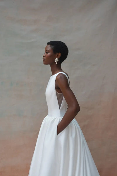 A person in the Audrey Wedding Dress by Jenny Yoo, featuring a white sleeveless design with pockets and an elegant boat neckline, standing sideways against a neutral background. The dress is available from Bergamot Bridal.