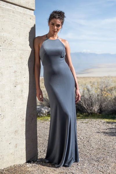 Woman in an elegant Bailey - Bridesmaid Dress by Jenny Yoo with a halter neckline posing beside a concrete structure with a natural landscape in the background from Bergamot Bridal.