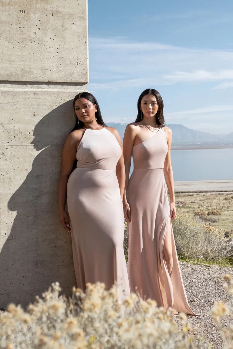 Two women posing in elegant beige stretch velvet gowns, one wearing the Cybill - Bridesmaid Dress by Jenny Yoo, against a concrete structure with a scenic backdrop from Bergamot Bridal.