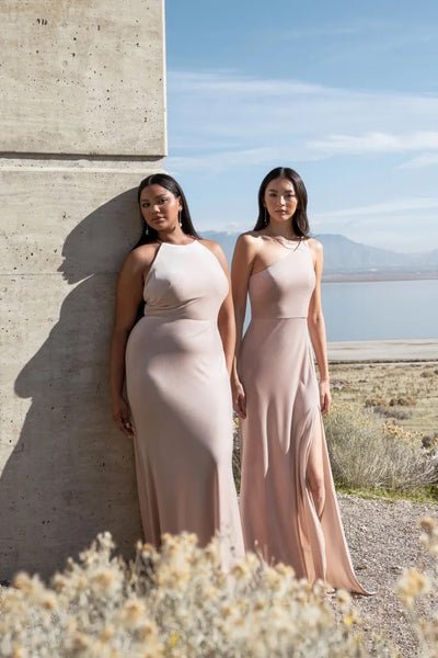 Two women posing in elegant beige stretch velvet gowns, one wearing the Cybill - Bridesmaid Dress by Jenny Yoo, against a concrete structure with a scenic backdrop from Bergamot Bridal.