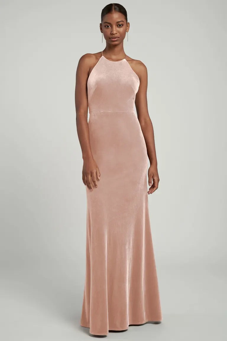 A woman standing in a blush-colored Bailey Bridesmaid Dress by Jenny Yoo with a halter neckline, ready for the bridal party from Bergamot Bridal.