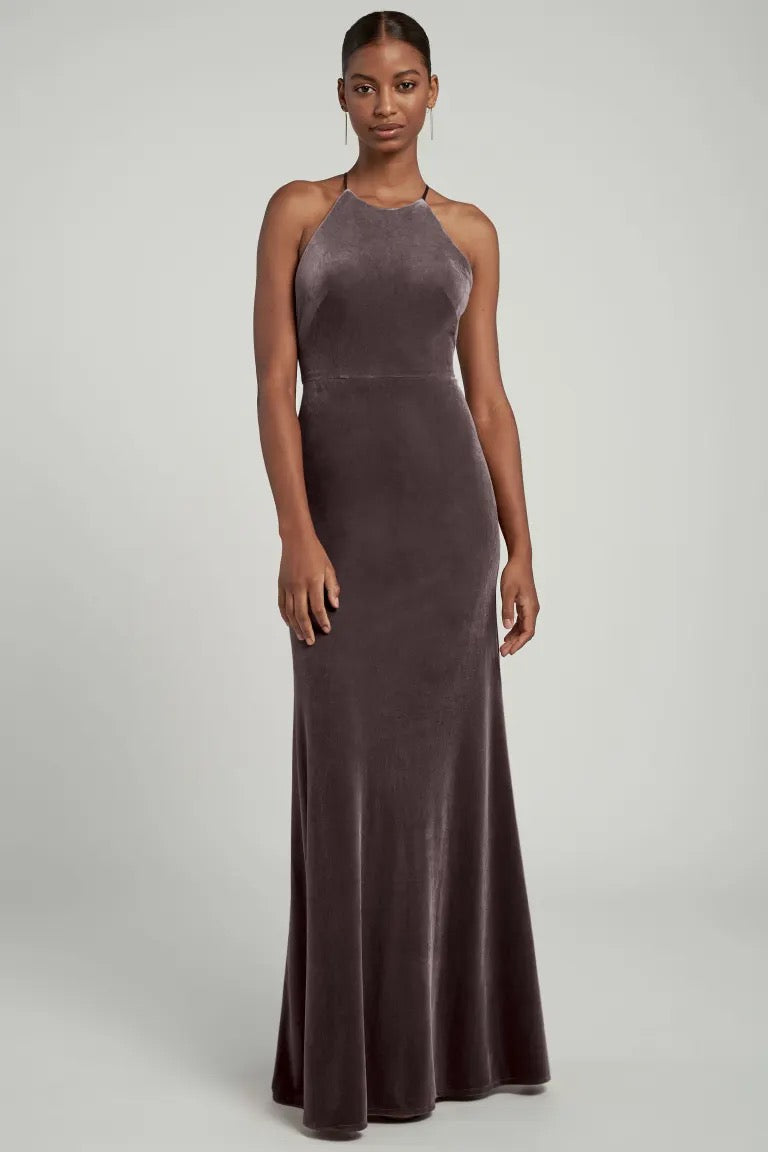 A woman wearing a sleeveless, halter neckline, full-length Bridesmaid Dress by Jenny Yoo in velvet fabric, posing against a plain background.