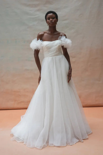 A woman stands wearing the Beatrice Shrug by Jenny Yoo, a flowing off-the-shoulder bridal gown with puffed sleeves crafted from pebbled organza, set against a minimalist backdrop.