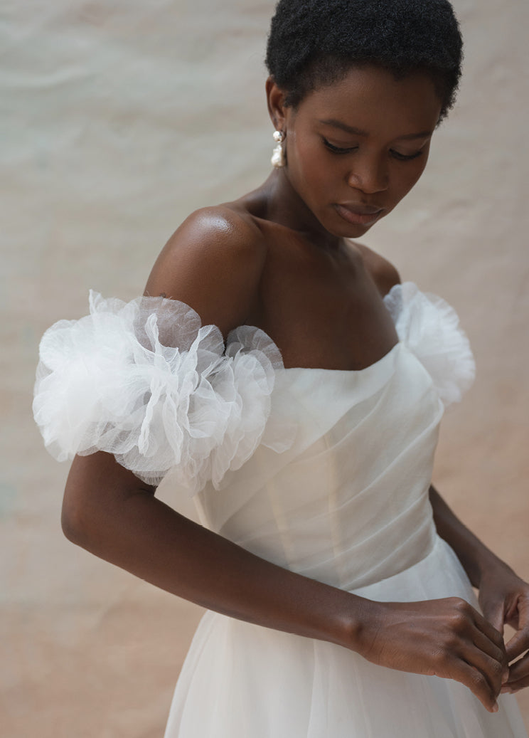 A person wearing the Beatrice Shrug by Jenny Yoo, from Bergamot Bridal, an off-shoulder white bridal gown with puffy tulle sleeves, looks down while standing against a neutral background.