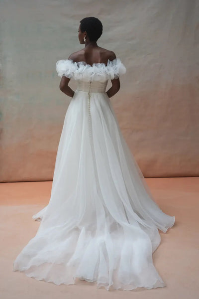 A person stands with their back to the camera, showcasing the Beatrice Shrug by Jenny Yoo from Bergamot Bridal, an off-the-shoulder white pebbled organza bridal gown featuring a long train and button details on the back. The backdrop consists of a gradient of soft brown tones.