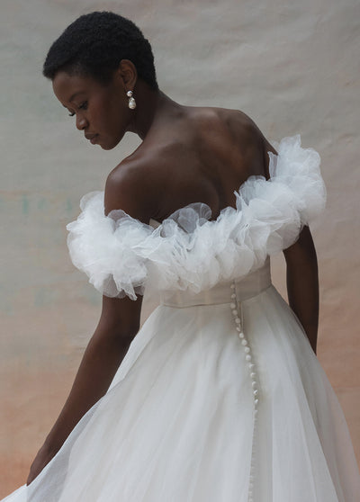 A woman with short hair and earrings is shown from the back wearing the 'Beatrice Shrug' by Jenny Yoo from Bergamot Bridal. The off-the-shoulder bridal gown features ruffled sleeves and a row of buttons down the back, made of elegant pebbled organza.