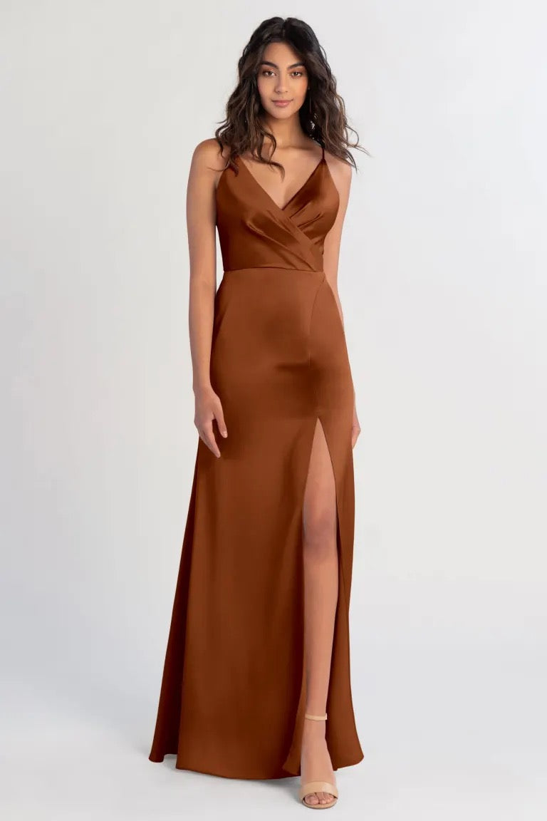A woman wearing a Beckette bridesmaid dress by Jenny Yoo in brown satin with a high slit stands poised against a gray background from Bergamot Bridal.