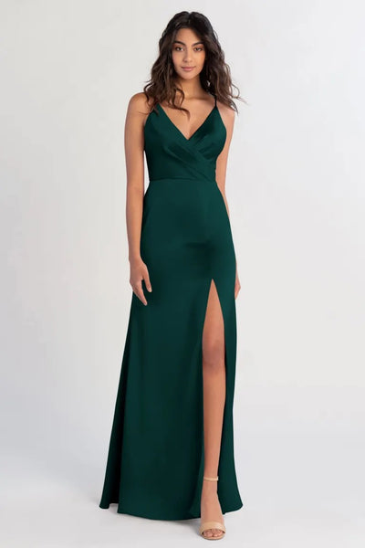 A woman wearing an elegant green satin Beckette Bridesmaid Dress by Jenny Yoo from Bergamot Bridal with a leg slit stands against a neutral background, exuding old Hollywood glamour.