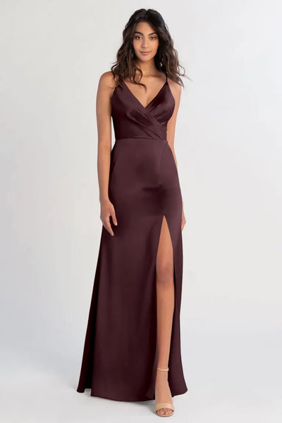 A woman in an elegant maroon satin Beckette bridesmaid dress by Jenny Yoo with a V-neck standing against a plain background from Bergamot Bridal.