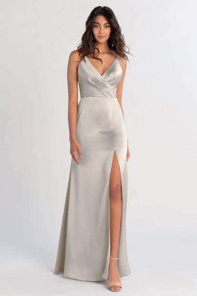Woman in a Beckette satin silver evening gown with a thigh-high slit and V-neck standing against a plain background by Bergamot Bridal.