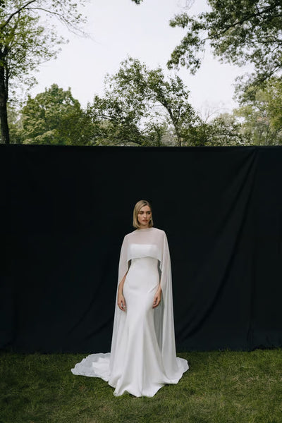 A woman wears the Belinda Cape by Jenny Yoo from Bergamot Bridal, standing on grass before a black backdrop outdoors, surrounded by trees.