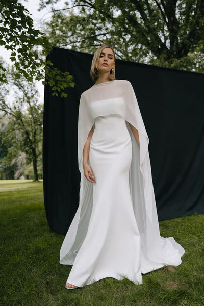 A woman in a fitted white bridal gown and the Belinda Cape by Jenny Yoo from Bergamot Bridal stands on grass before a black backdrop, with trees behind her.