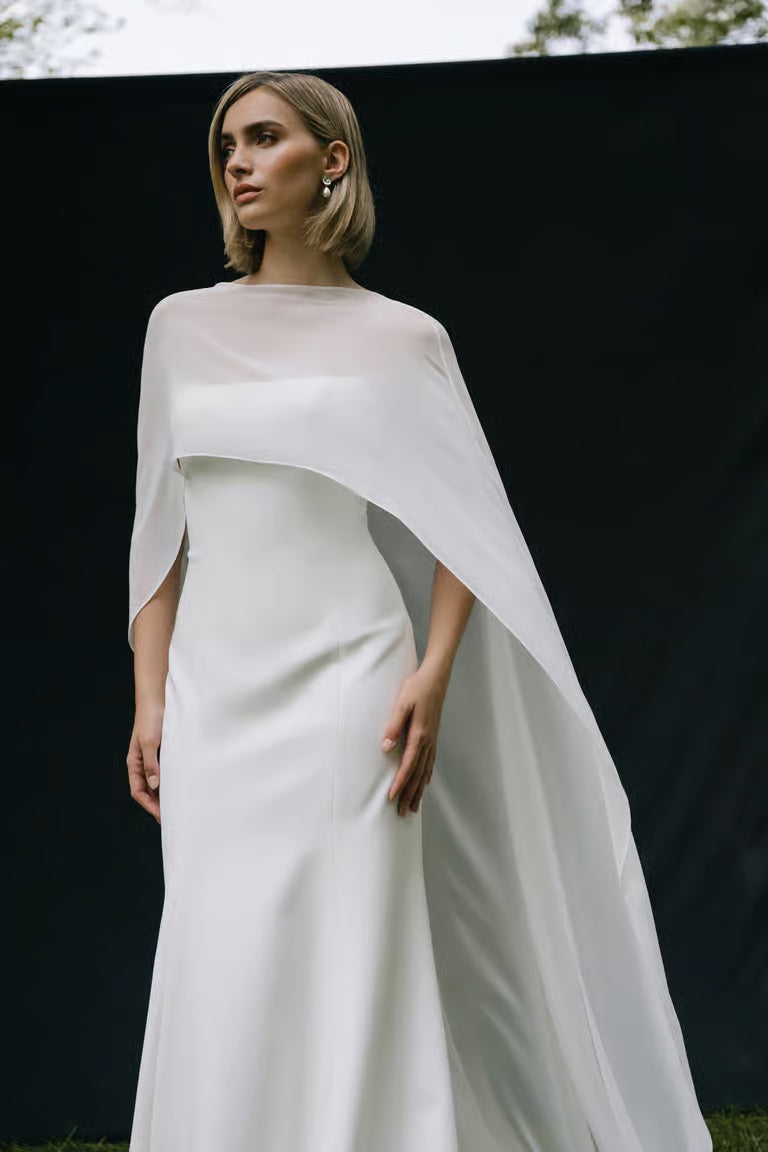 A woman stands against a dark backdrop, looking to the side in a sleek white bridal gown styled with the Belinda Cape by Jenny Yoo from Bergamot Bridal.
