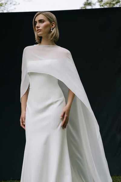 A woman stands against a dark backdrop, looking to the side in a sleek white bridal gown styled with the Belinda Cape by Jenny Yoo from Bergamot Bridal.