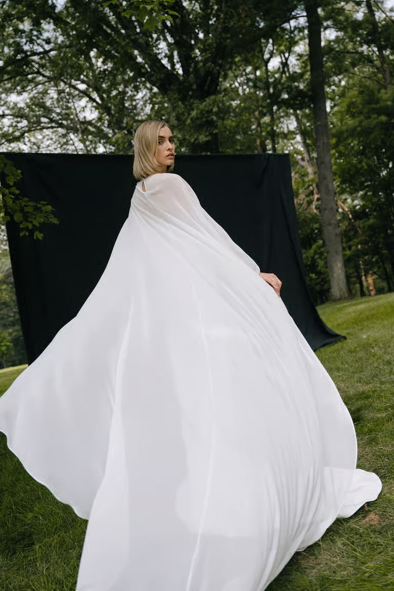 A woman wears the Belinda Cape by Jenny Yoo from Bergamot Bridal, standing outdoors on grass with a black backdrop and trees behind her.