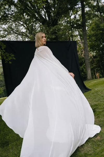A woman wears the Belinda Cape by Jenny Yoo from Bergamot Bridal, standing outdoors on grass with a black backdrop and trees behind her.
