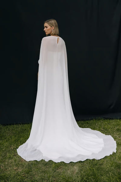A woman stands on grass facing away, wearing the Belinda Cape by Jenny Yoo from Bergamot Bridal—a long, flowing white wedding cape—set against a dark backdrop.