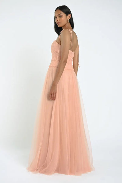 A woman stands facing slightly away, wearing the elegant Berkley Bridesmaid Dress by Jenny Yoo from Bergamot Bridal, crafted from soft tulle fabric in a long, peach-colored design. The dress features thin shoulder straps and a fitted bodice that exudes a romantic allure.