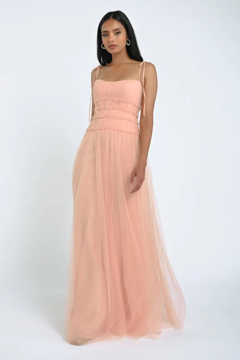 A woman wearing a long, romantic Berkley Bridesmaid Dress by Jenny Yoo in pink, made of soft tulle fabric with thin straps from Bergamot Bridal stands against a plain white background.