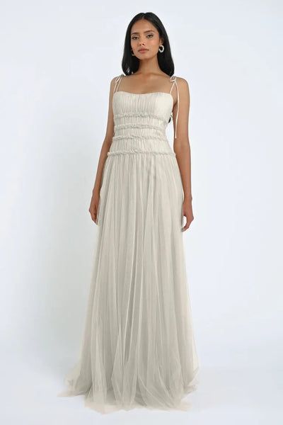 A woman stands in a romantic, light gray, floor-length Berkley Bridesmaid Dress by Jenny Yoo from Bergamot Bridal, made of soft tulle fabric, against a plain white background.