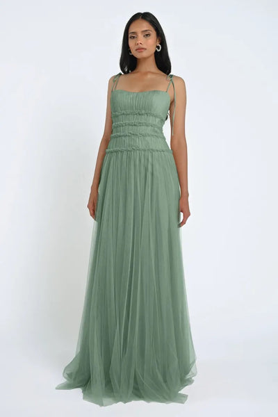 A woman with long dark hair is standing against a plain white background, wearing the Berkley Bridesmaid Dress by Jenny Yoo from Bergamot Bridal. The floor-length gown, in a romantic green hue, is sleeveless and made from soft tulle fabric. She looks directly at the camera.