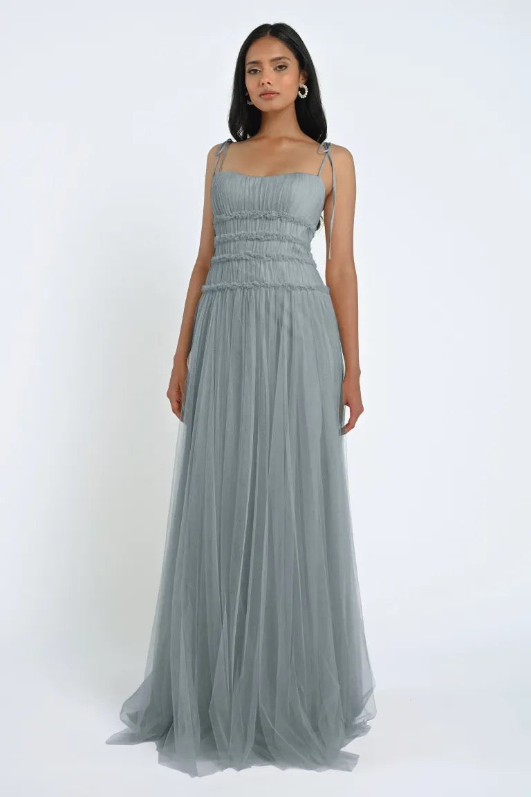 A woman stands wearing the Berkley Bridesmaid Dress by Jenny Yoo from Bergamot Bridal. The floor-length, sleeveless dress features ruched detailing and thin shoulder straps, crafted from soft tulle fabric. She is looking directly ahead against a plain white background, exuding a romantic elegance.