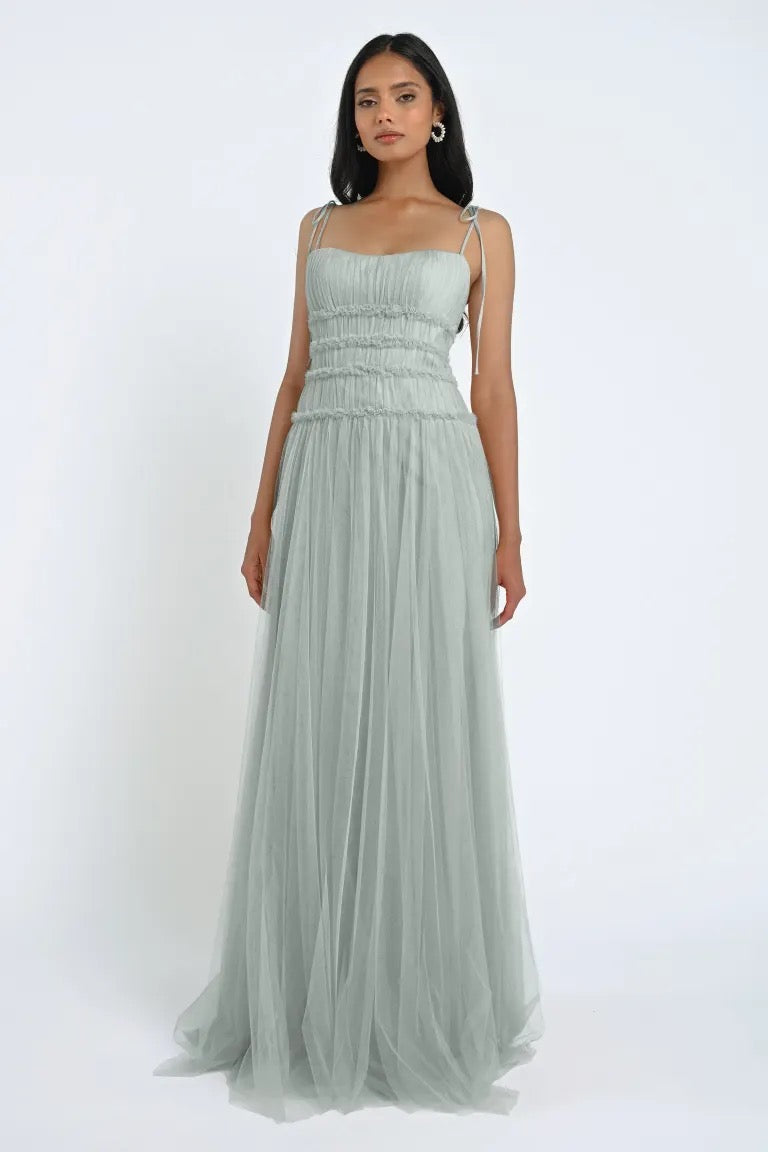 A woman is standing against a plain background, wearing the romantic Berkley Bridesmaid Dress by Jenny Yoo from Bergamot Bridal, a sleeveless floor-length light grey gown with delicate straps and gathered detailing at the bodice, crafted from soft tulle fabric.