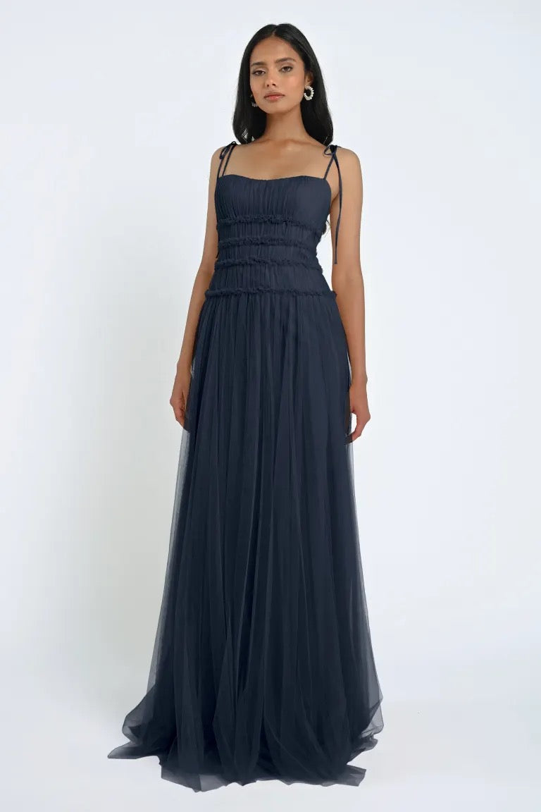 A woman stands against a plain background wearing the romantic Berkley Bridesmaid Dress by Jenny Yoo from Bergamot Bridal, a navy blue floor-length gown featuring spaghetti straps and a gathered bodice made from soft tulle fabric.