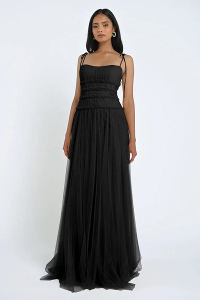 A woman stands wearing the Berkley Bridesmaid Dress by Jenny Yoo from Bergamot Bridal, a long, black dress featuring a fitted bodice and a flowing skirt made of soft tulle fabric. With her straight, dark hair and minimal accessories set against a plain white background, she exudes romantic elegance.