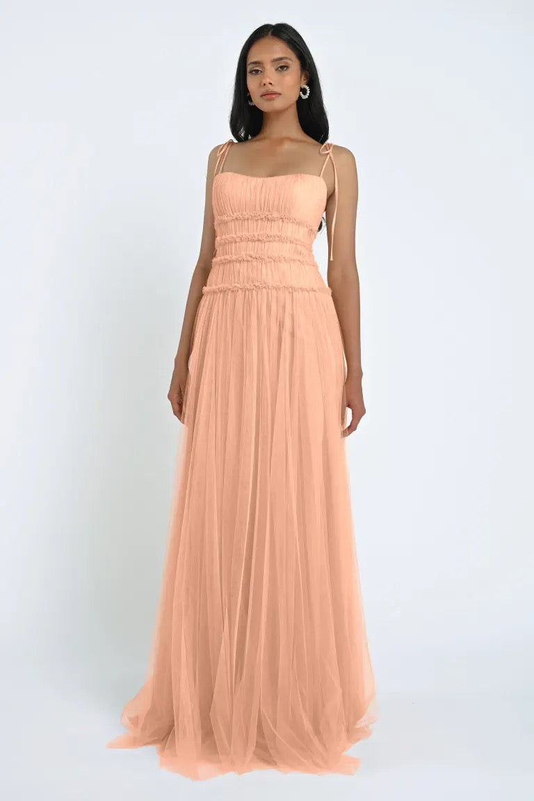 A woman wearing a romantic Berkley Bridesmaid Dress by Jenny Yoo from Bergamot Bridal, featuring a long, peach-colored design with thin straps and a smocked bodice made from soft tulle fabric, stands against a plain white background.