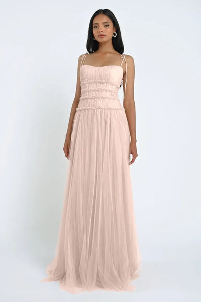 A woman stands wearing the romantic, light pink Berkley Bridesmaid Dress by Jenny Yoo from Bergamot Bridal. The dress features shoulder straps and a gathered bodice, with soft tulle fabric flowing gracefully as she faces forward against a plain white background.