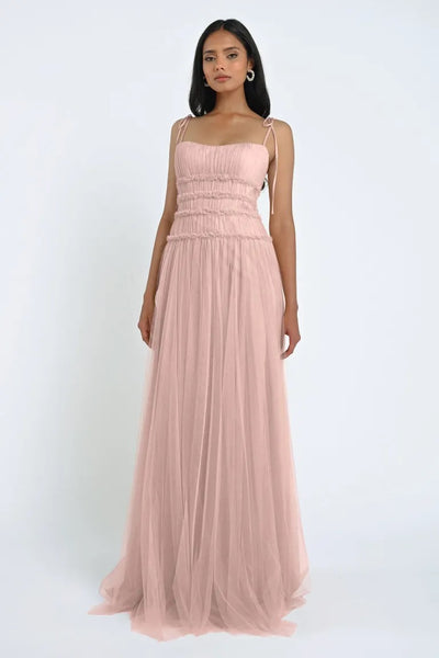 A woman is wearing a long, pink, sleeveless Berkley Bridesmaid Dress by Jenny Yoo from Bergamot Bridal. The romantic gown has a fitted, ruched bodice and thin straps, and it features soft tulle fabric in the flowing, semi-transparent skirt as she stands against a plain white background.