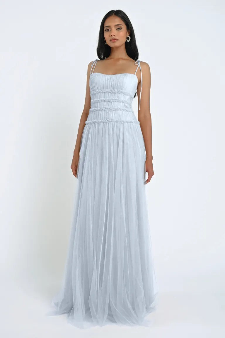 A woman stands against a white background wearing the Berkley Bridesmaid Dress by Jenny Yoo, a light blue, floor-length gown from Bergamot Bridal. This dress features thin straps and a gathered waist, made of soft tulle fabric that exudes a romantic charm.