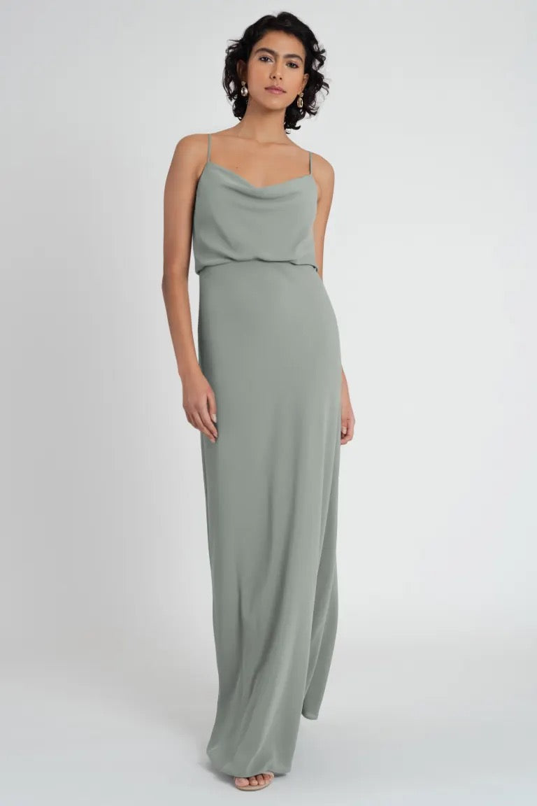 A woman in a simple, yet elegant sage green Bianca bridesmaid dress by Jenny Yoo stands against a white background.