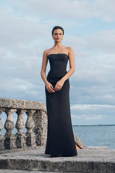 A woman in an elegant black Paige - Jenny Yoo Bridesmaid Dress made from Luxe Faille fabric stands by a stone balustrade against a backdrop of a calm sea and sky.