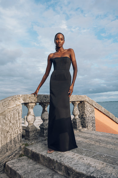 A woman in an elegant Jenny Yoo Bridesmaid Dress with a fit and flare skirt, standing by a stone balustrade with the ocean in the background from Bergamot Bridal.