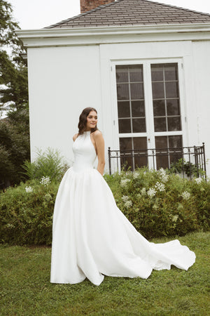 A woman in the sleeveless white Blake gown by Jenny Yoo from Bergamot Bridal stands on grass before a white building with large windows and lush greenery.