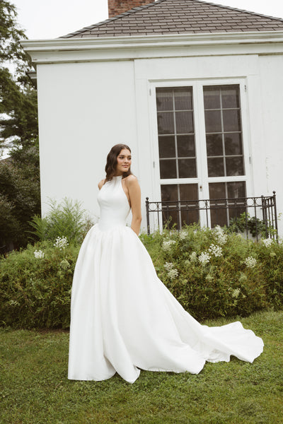A woman in the sleeveless white Blake gown by Jenny Yoo from Bergamot Bridal stands on grass before a white building with large windows and lush greenery.