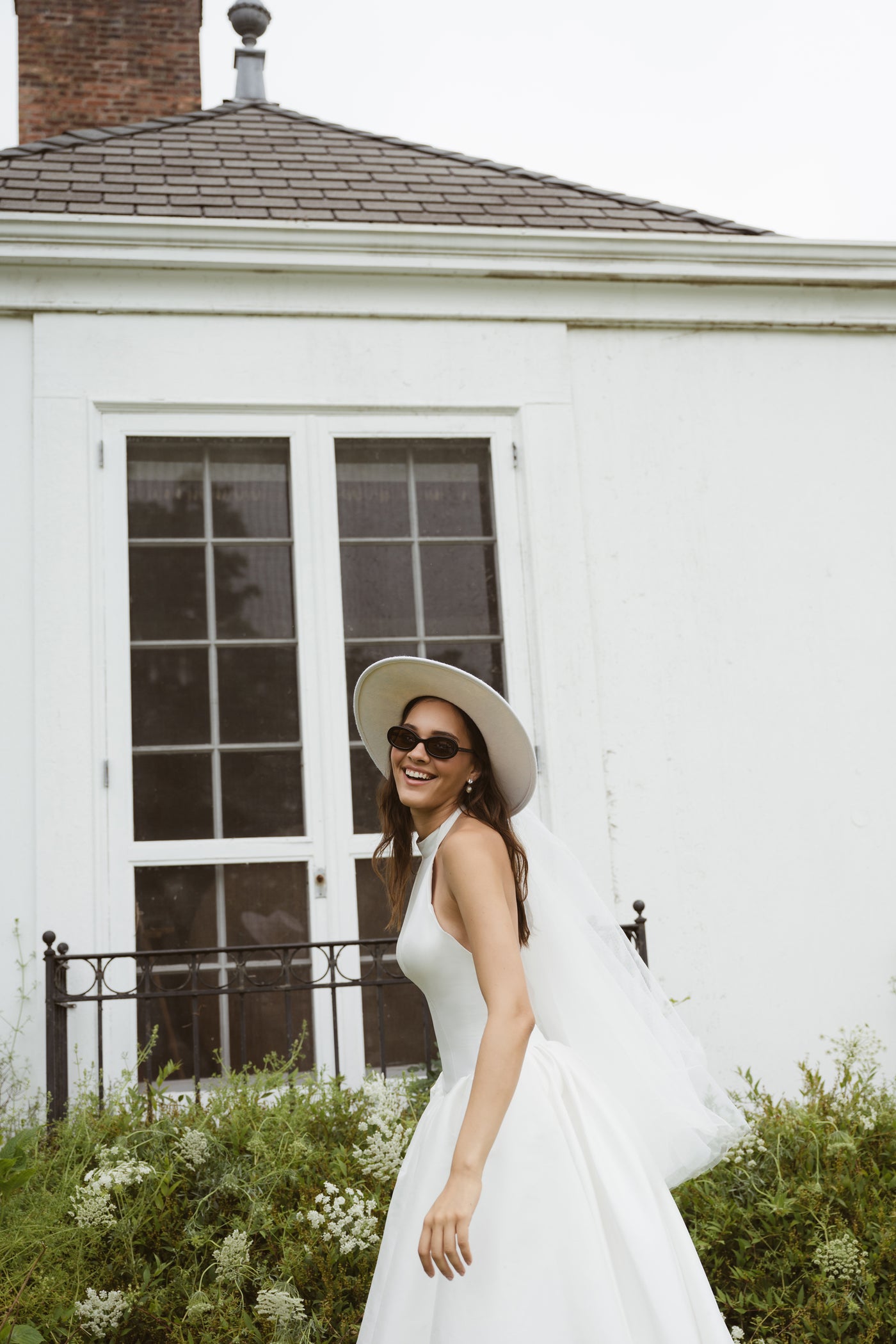 A woman smiles outside a white building, wearing the Blake by Jenny Yoo gown from Bergamot Bridal—a ball skirt dress paired with a hat and sunglasses.