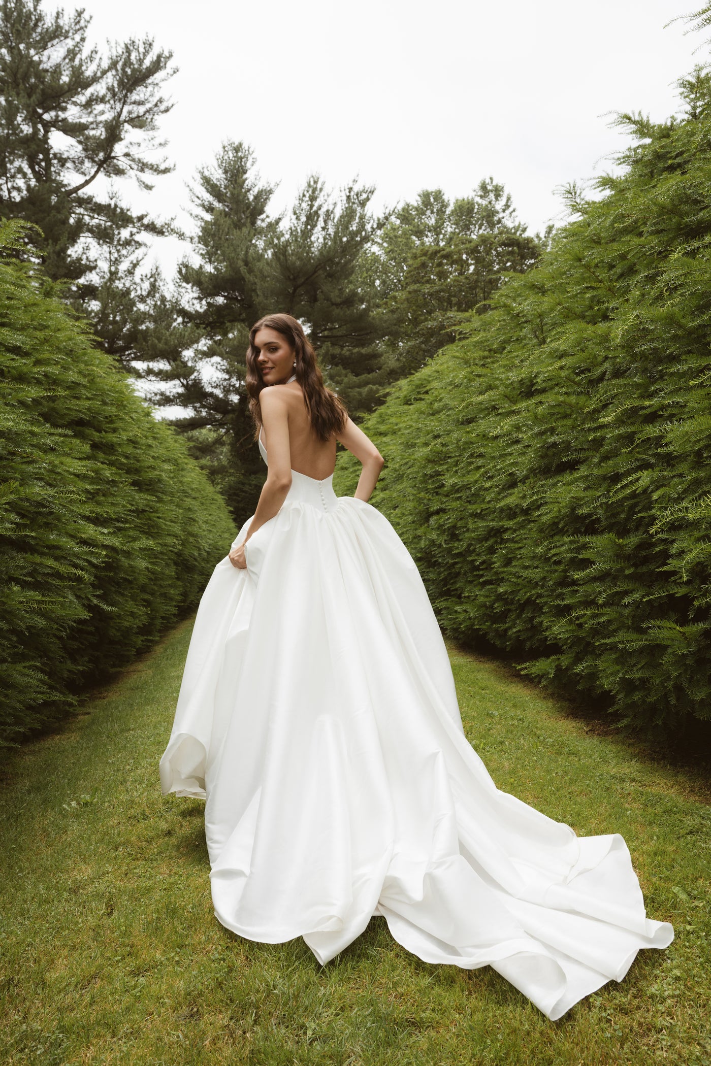 A woman in the Blake by Jenny Yoo gown from Bergamot Bridal, featuring a ball skirt, stands on a grassy path between tall green hedges, gazing over her shoulder.