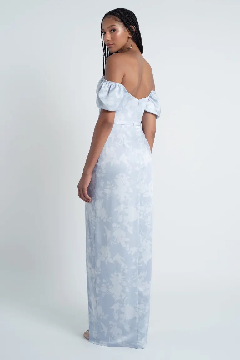 A woman wearing an off-shoulder, floor-length Jenny Yoo Bridesmaid Dress with a Corsage printed satin pattern, standing side-on to the camera from Bergamot Bridal.