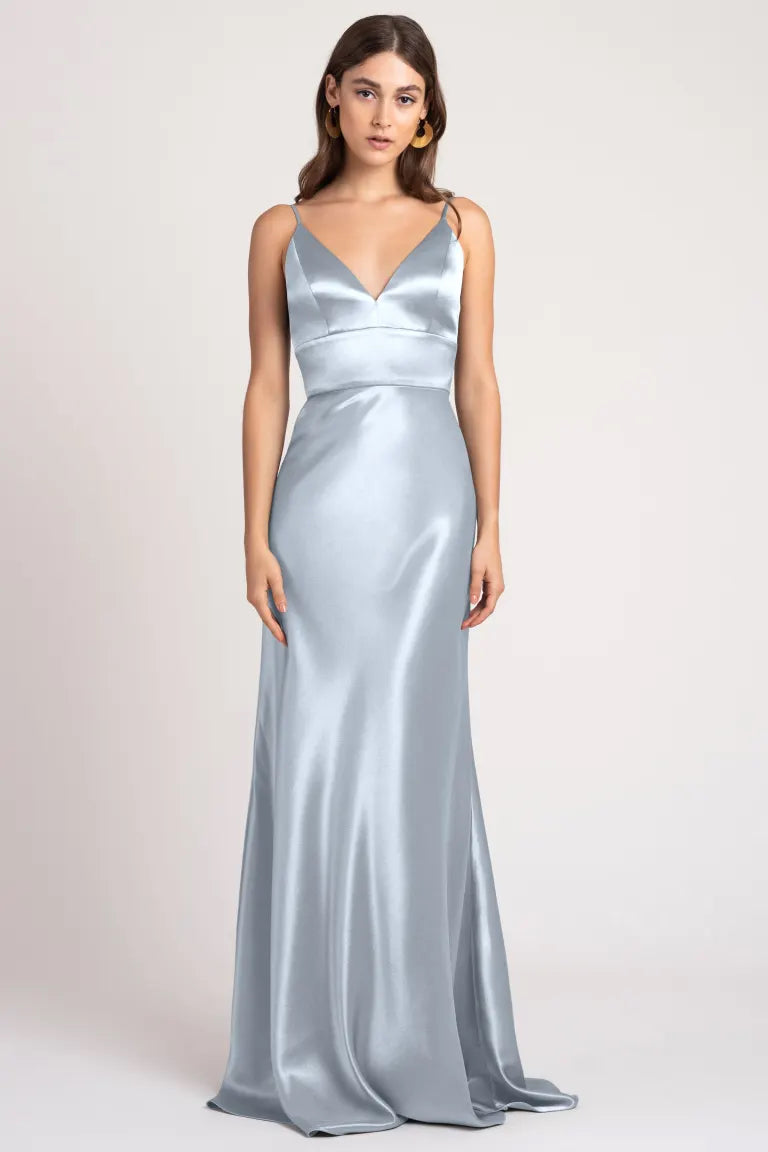 A woman is standing in a silver satin back crepe Brenna - Bridesmaid Dress by Jenny Yoo from Bergamot Bridal, featuring thin straps and a V-neckline. She has long hair and a neutral expression, set against a plain background.