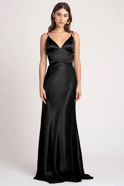 A woman in an elegant black Brenna dress by Jenny Yoo, made of satin back crepe fabric, standing against a neutral background.